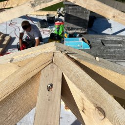 T&M BUDOWNICTWO SPÓŁKA Z OGRANICZONĄ ODPOWIEDZIALNOŚCIĄ - Wiązar dachowy z jasnego drewna w trakcie montażu. W tle murarz przy pracy, narzędzia i materiały budowlane. Widok z góry na konstrukcję dachu.