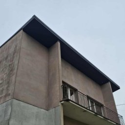 T&M BUDOWNICTWO SPÓŁKA Z OGRANICZONĄ ODPOWIEDZIALNOŚCIĄ - Dwukondygnacyjny budynek z balkonem i płaskim dachem, widziany z dołu pod kątem. Ściany w odcieniach beżu i szarości, obok drzewo iglaste na tle pochmurnego nieba.