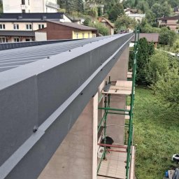 T&M BUDOWNICTWO SPÓŁKA Z OGRANICZONĄ ODPOWIEDZIALNOŚCIĄ - Szary dach z metalu z widocznymi elementami mocującymi, rusztowanie obok budynku, zieleń w tle. Ujęcie z góry.