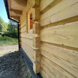 Fragment ściany zewnętrznej domu z bali, widoczny detal łączenia bali na rogu budynku, z czarną folią fundamentową przy ziemi. Surowe drewno, widoczne słoje.