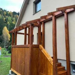 Drewniana altana z zadaszeniem, przylegająca do budynku z beżową elewacją, schody wejściowe. Na trawie narzędzia. Widok na las w tle.