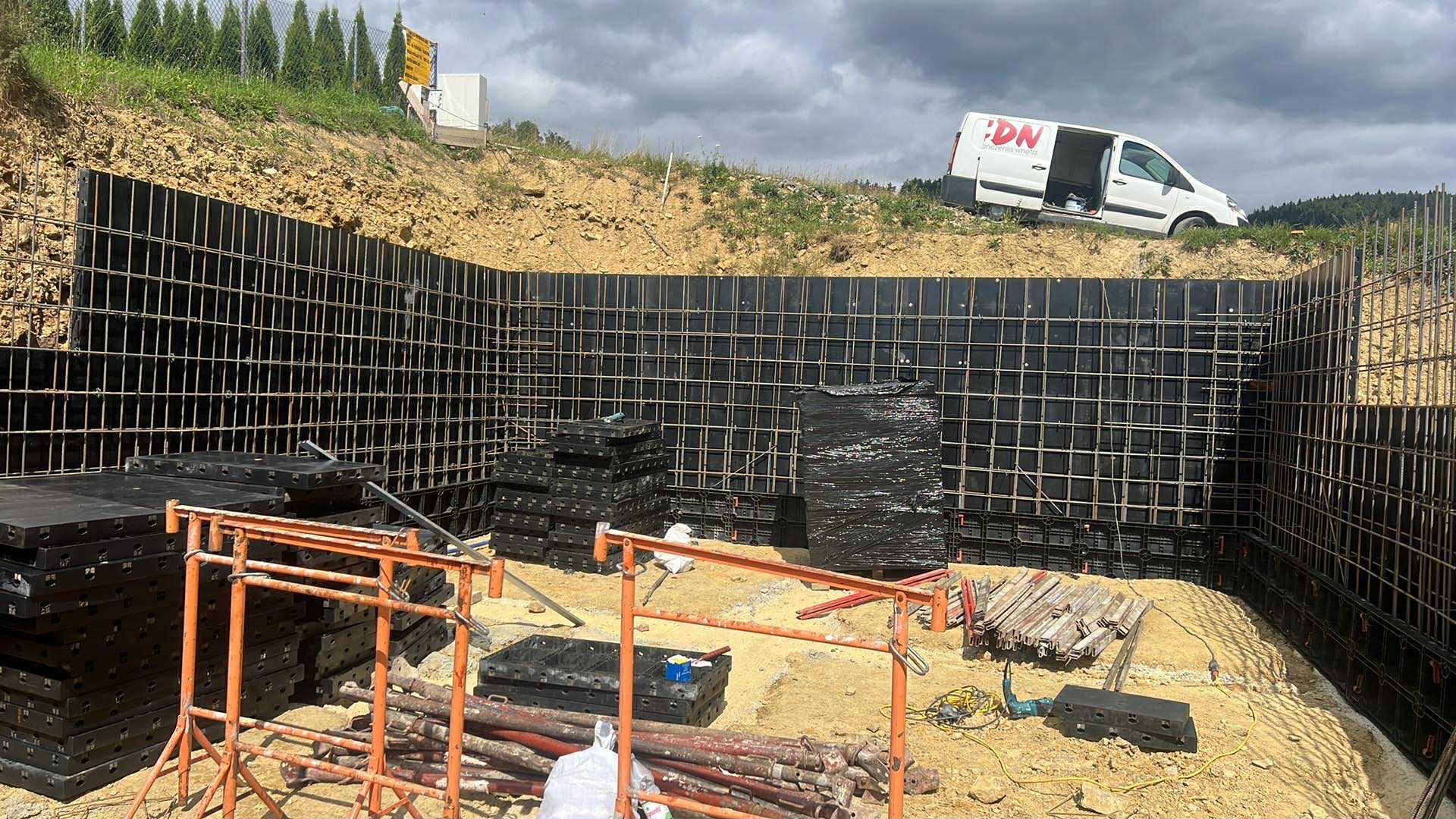 Zbrojenie i szalunek pod fundamenty w Powroźniku. Widoczne metalowe pręty i czarne płyty szalunkowe. Na górze skarpy zaparkowany biały samochód dostawczy.