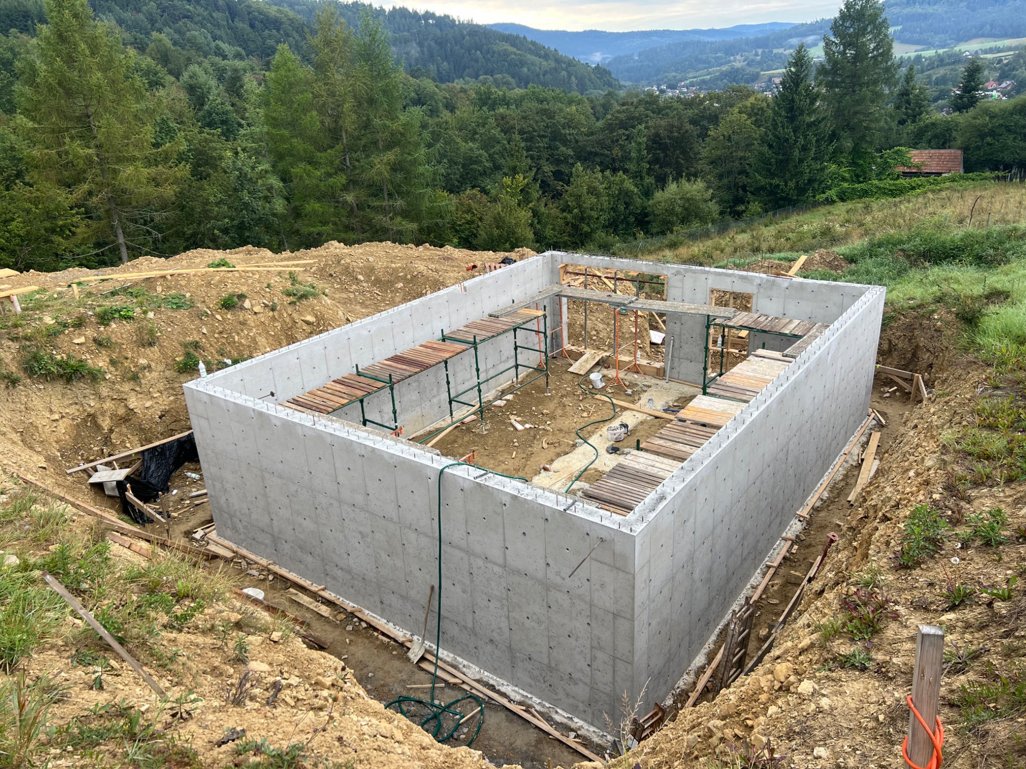 Wylany fundament pod budynek na tle zieleni i wzgórz. Widoczne szalunki i zbrojenia. Surowy betonowy fundament w wykopie, przygotowany do dalszych prac budowlanych.
