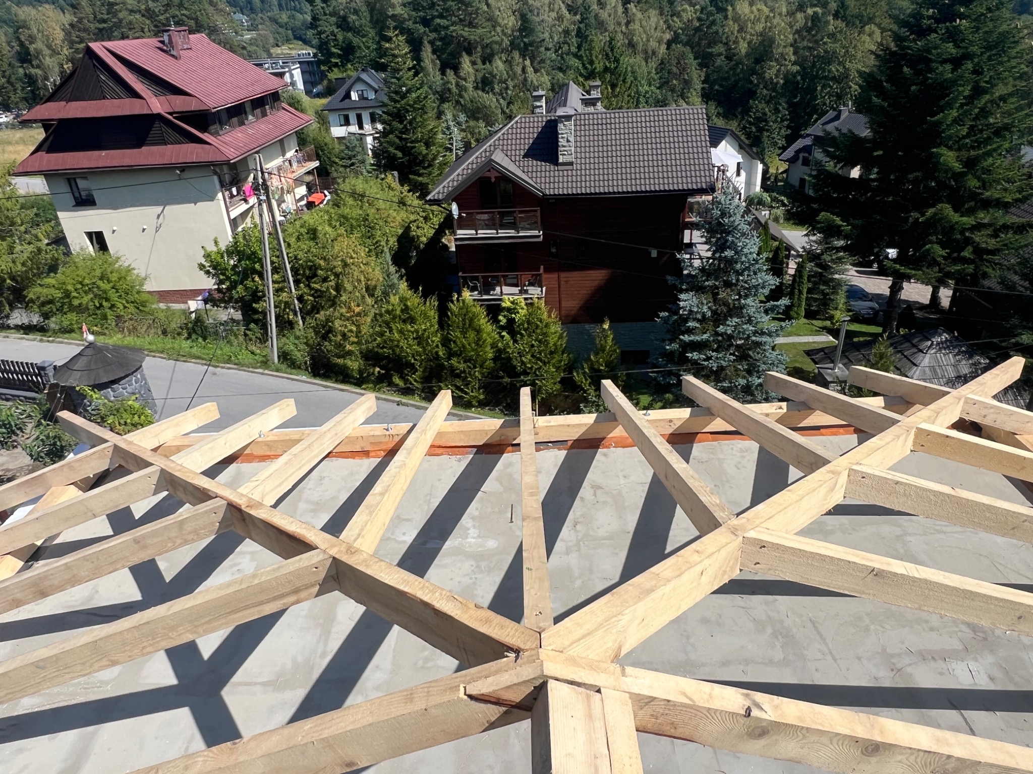 Wiązar dachowy z jasnego drewna na betonowej powierzchni, w tle zabudowa domów i las. Widok z góry, oświetlenie słoneczne.