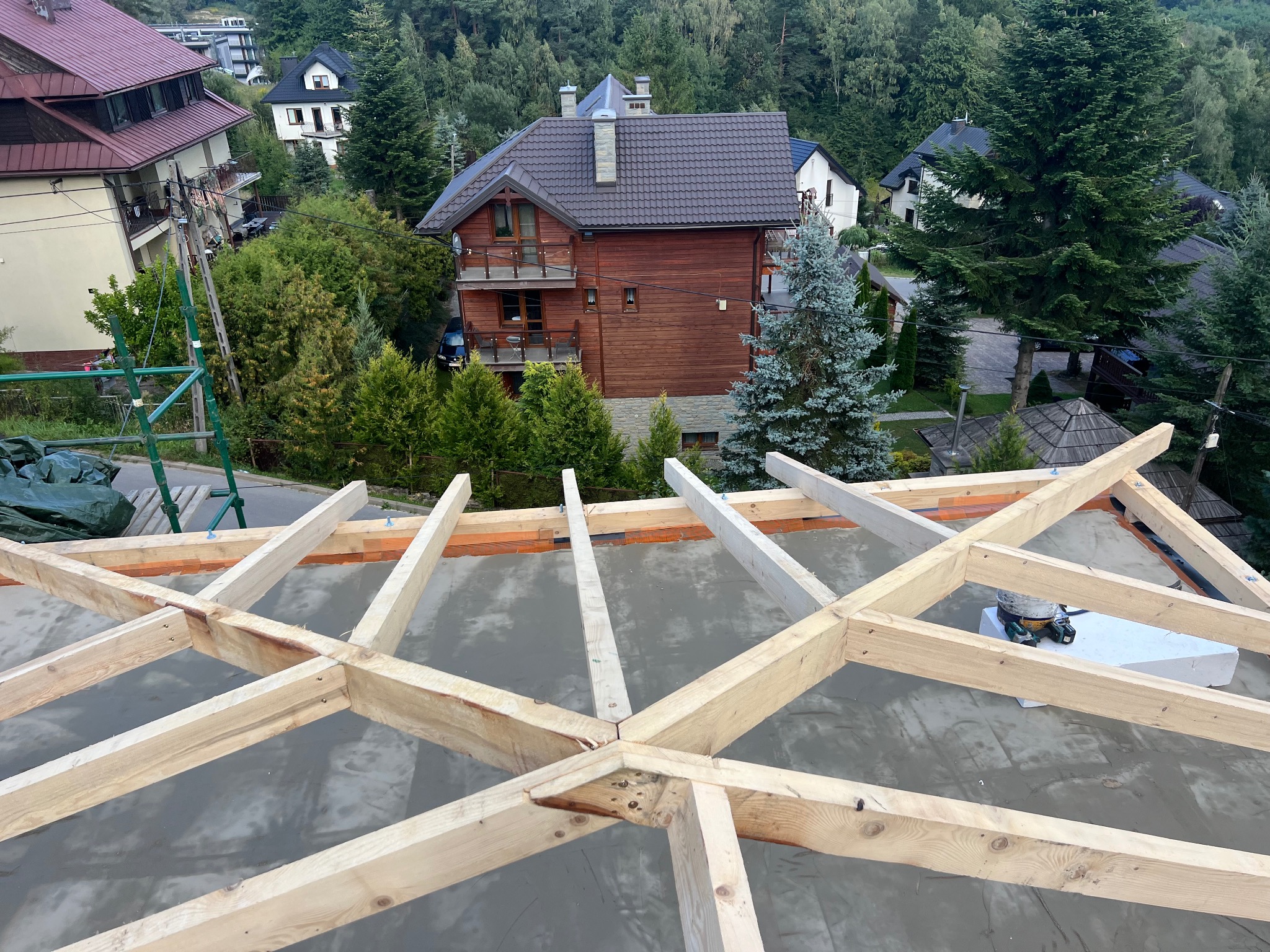 Wznoszenie drewnianej konstrukcji dachu na tle zabudowy jednorodzinnej i zieleni. Widoczna szara powierzchnia dachu i elementy drewniane więźby.