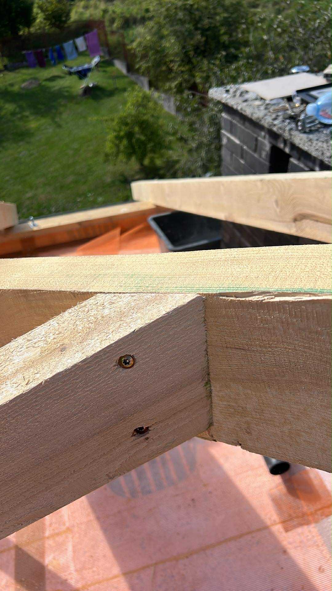 Fragment drewnianej konstrukcji dachu z widocznym wkrętem, membrana dachowa w tle, zieleń i fragment budynku w górnej części kadru.