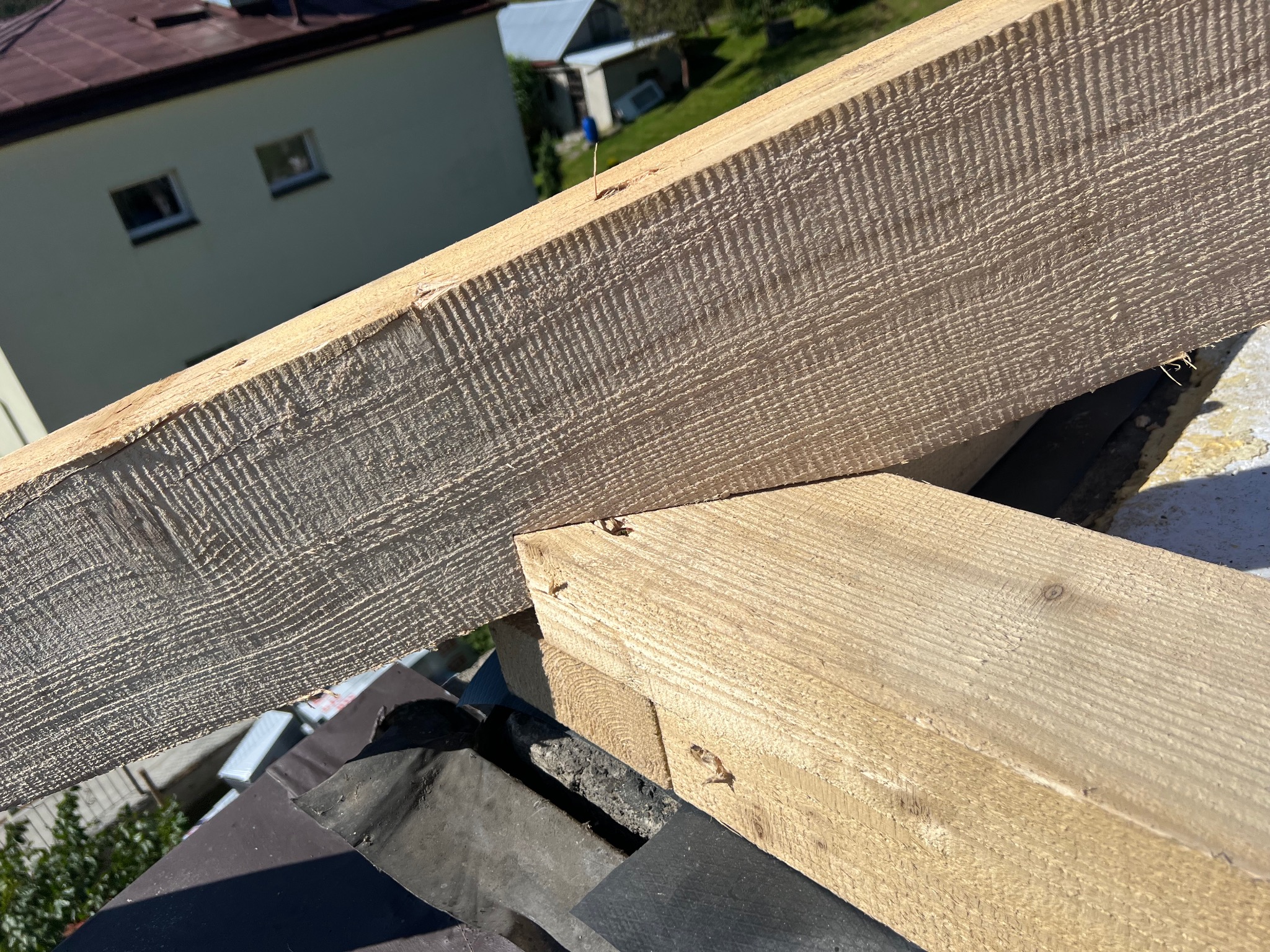 Fragment drewnianej więźby dachowej z widocznymi elementami konstrukcyjnymi i gwoździami na tle budynku i zieleni. Kąt ujęcia z góry, ostre światło.