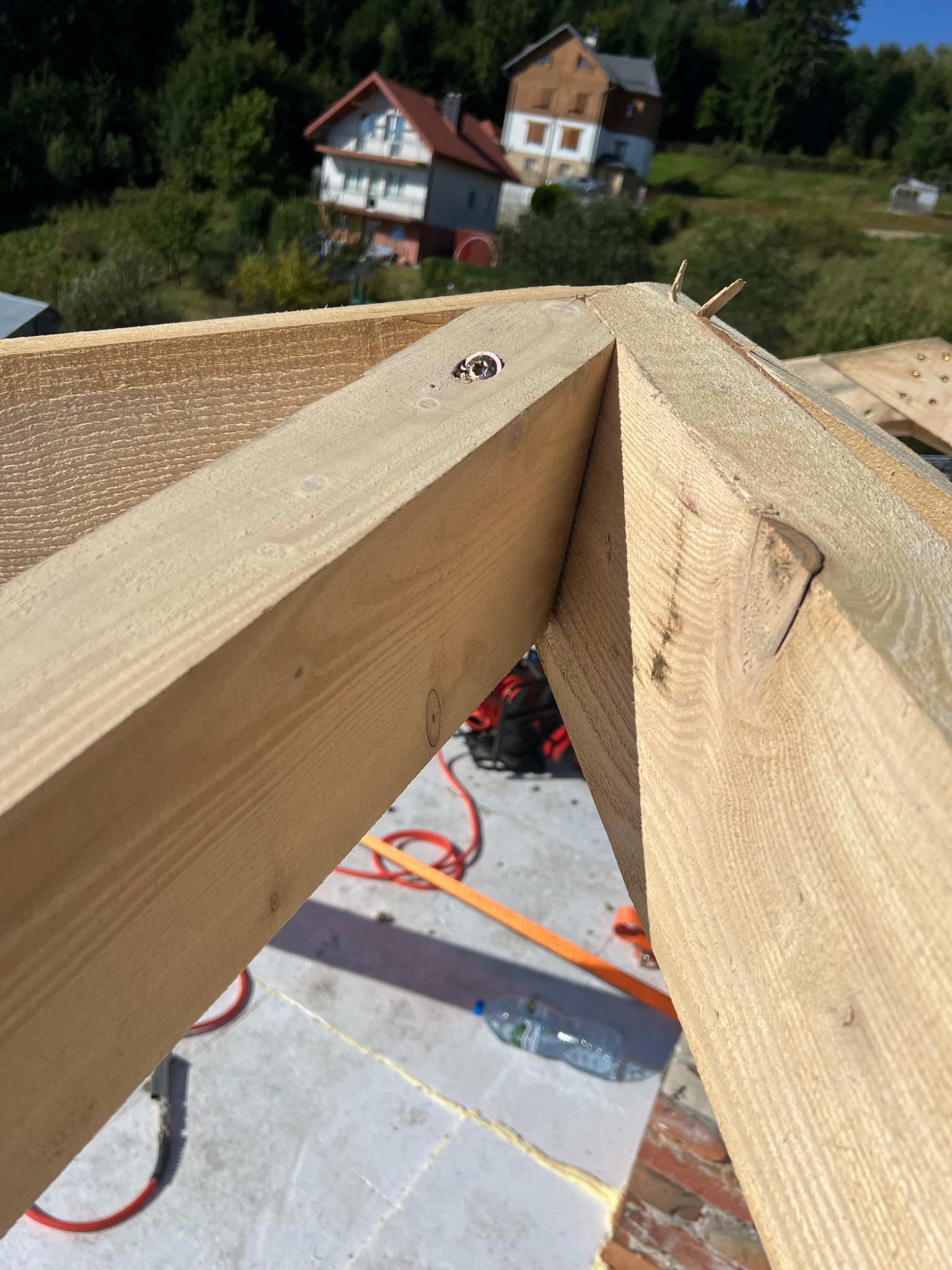 Fragment drewnianej więźby dachowej w trakcie montażu, z widocznym złączeniem elementów i narzędziami w tle, na tle zieleni i zabudowań.