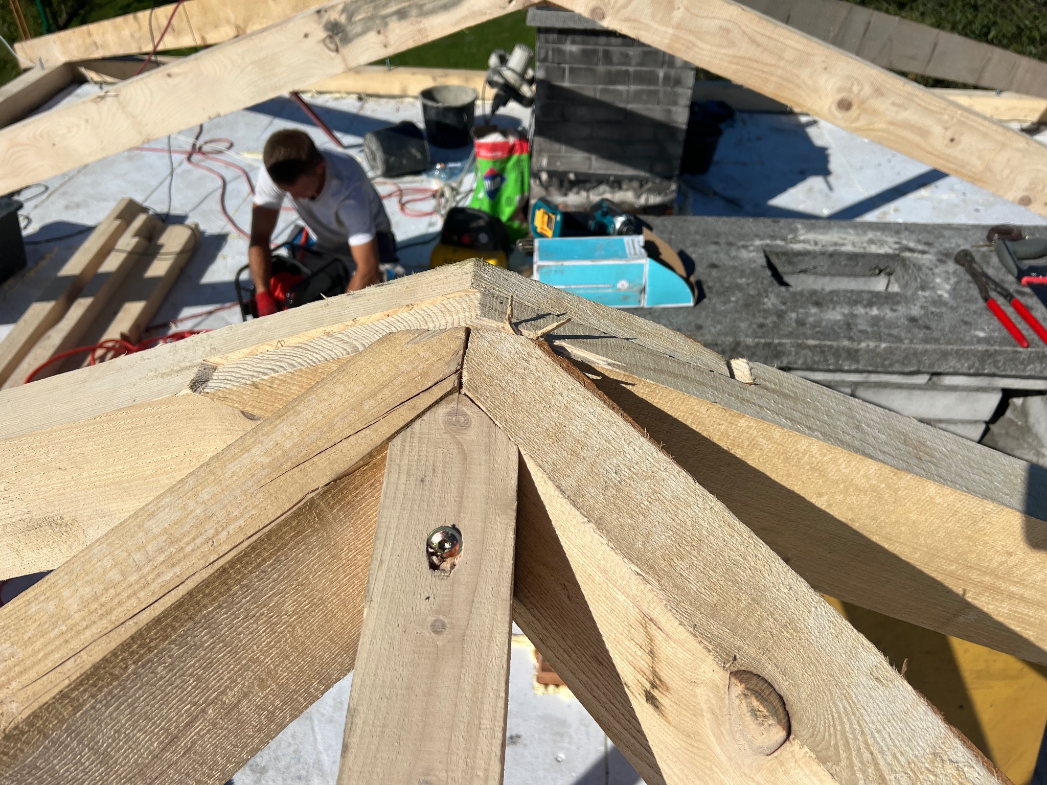 Wiązar dachowy z jasnego drewna w trakcie montażu. W tle murarz przy pracy, narzędzia i materiały budowlane. Widok z góry na konstrukcję dachu.