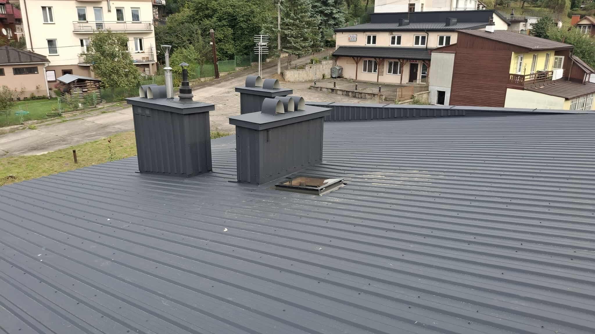 Szary, blaszany dach z trzema kominami i świetlikiem. Widok z góry na budynki w tle. Nowoczesne wykończenie.