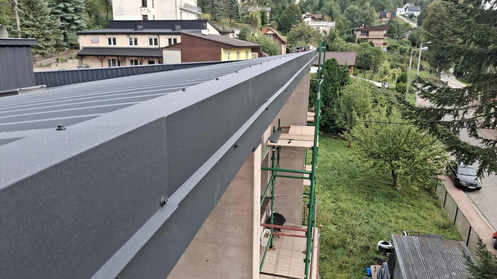 Szary, metalowy dach płaski z widocznymi mocowaniami, fragment rusztowania, budynki w tle. Ujęcie z góry.