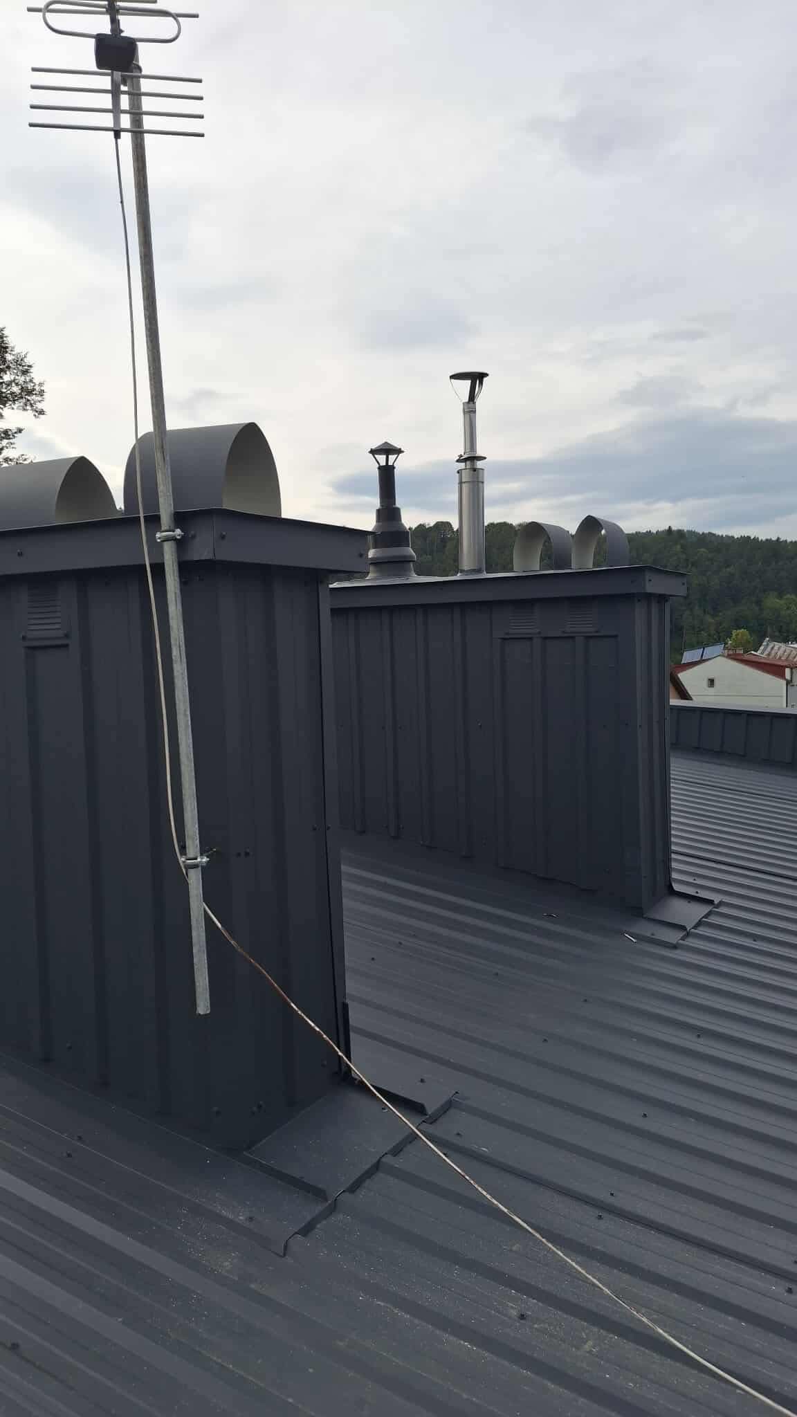 Szary, blaszany dach z kominami wentylacyjnymi i anteną telewizyjną. Widok na okoliczne budynki i zalesione wzgórza w tle. Pochmurne niebo.