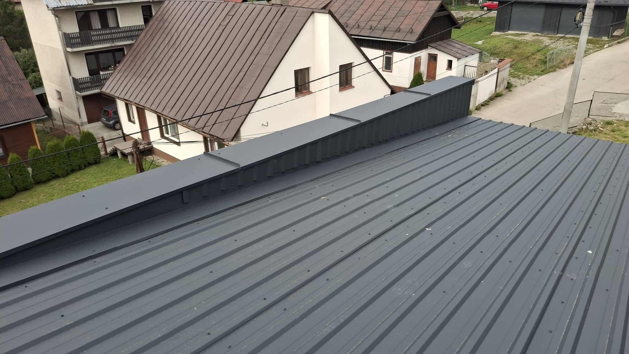 Szary, blaszany dach z widocznymi śrubami i elementami wykończeniowymi na tle zabudowy mieszkalnej. Ujęcie z góry.