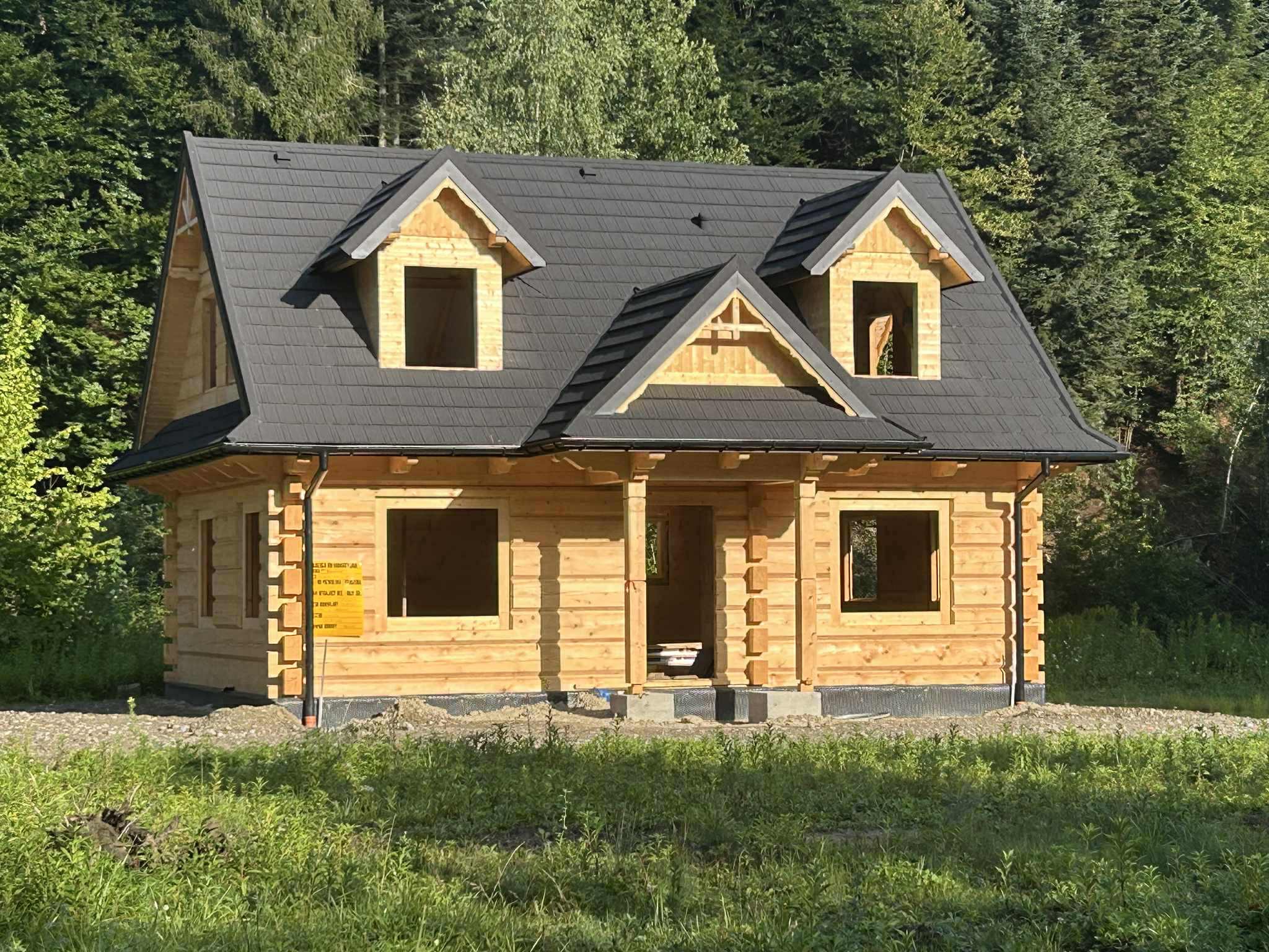 Surowy stan domu z bali z ciemnym dachem na tle zielonego lasu. Widoczne niezabudowane otwory okienne i drzwiowe. Naturalne oświetlenie, trawa w pierwszym planie.