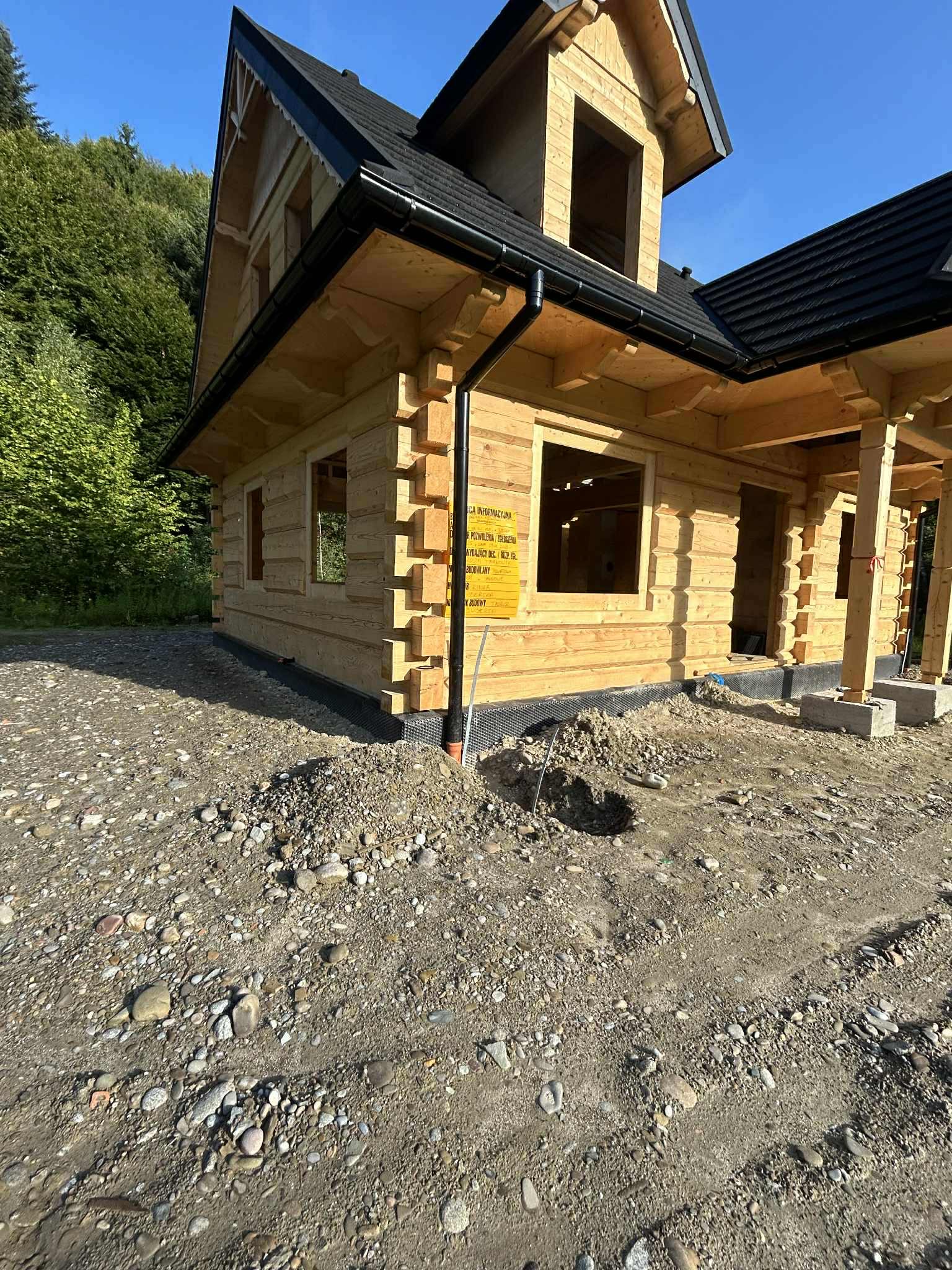 Surowy dom z bali z czarnym dachem, w trakcie budowy, na tle zieleni. Widoczne elementy konstrukcyjne i tablica informacyjna. Ujęcie w słoneczny dzień.