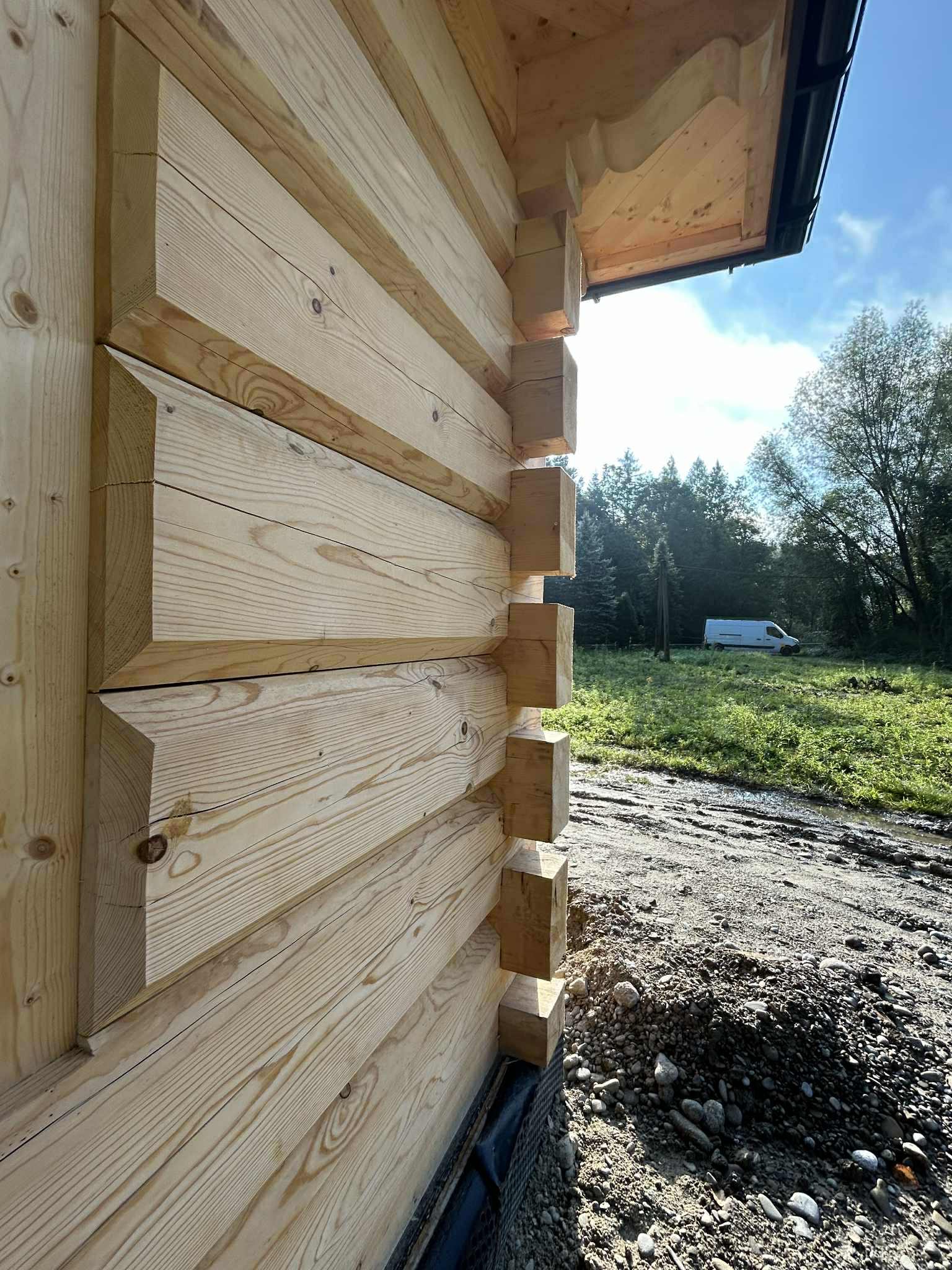 Fragment ściany domu z bali z widocznymi detalami łączeń, surowe drewno, w tle zieleń i błękit nieba. Ujęcie z dołu podkreśla solidną konstrukcję.