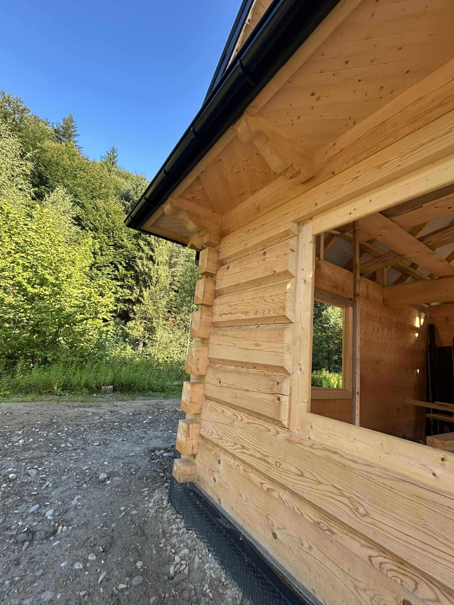 Surowy stan domu z bali w trakcie budowy, widoczne drewniane ściany z oknem bez ramy i fragment dachu z czarną rynną na tle zieleni.