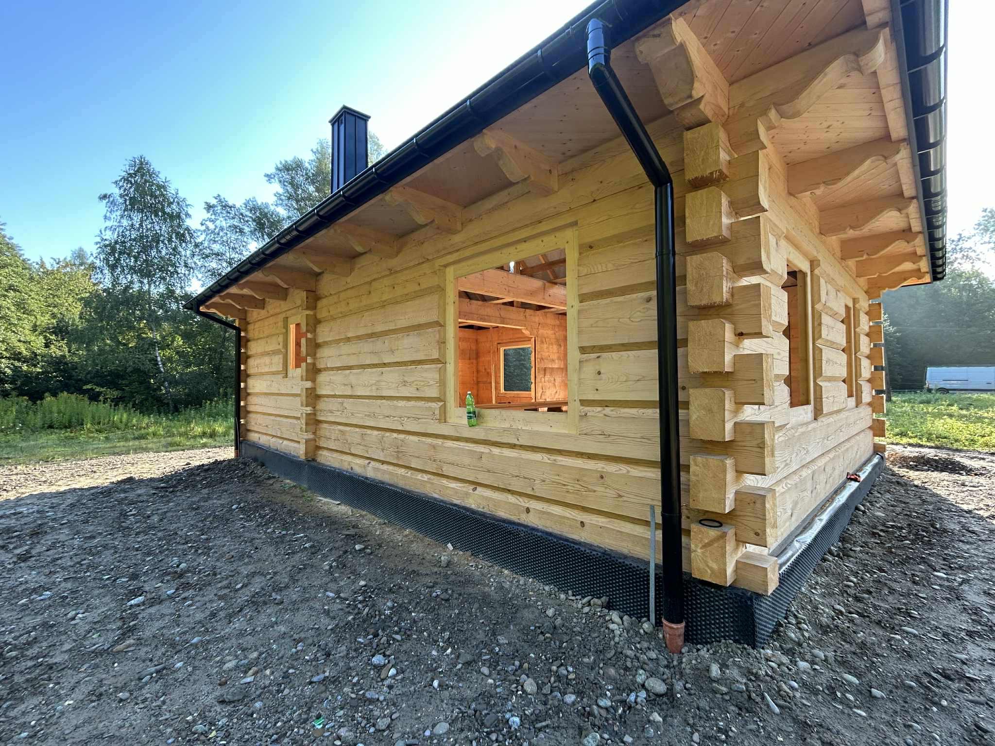 Surowy dom z bali w trakcie budowy, widoczne drewniane ściany, okno bez szyb, czarny komin i system rynnowy. Widok z zewnątrz na plac budowy.