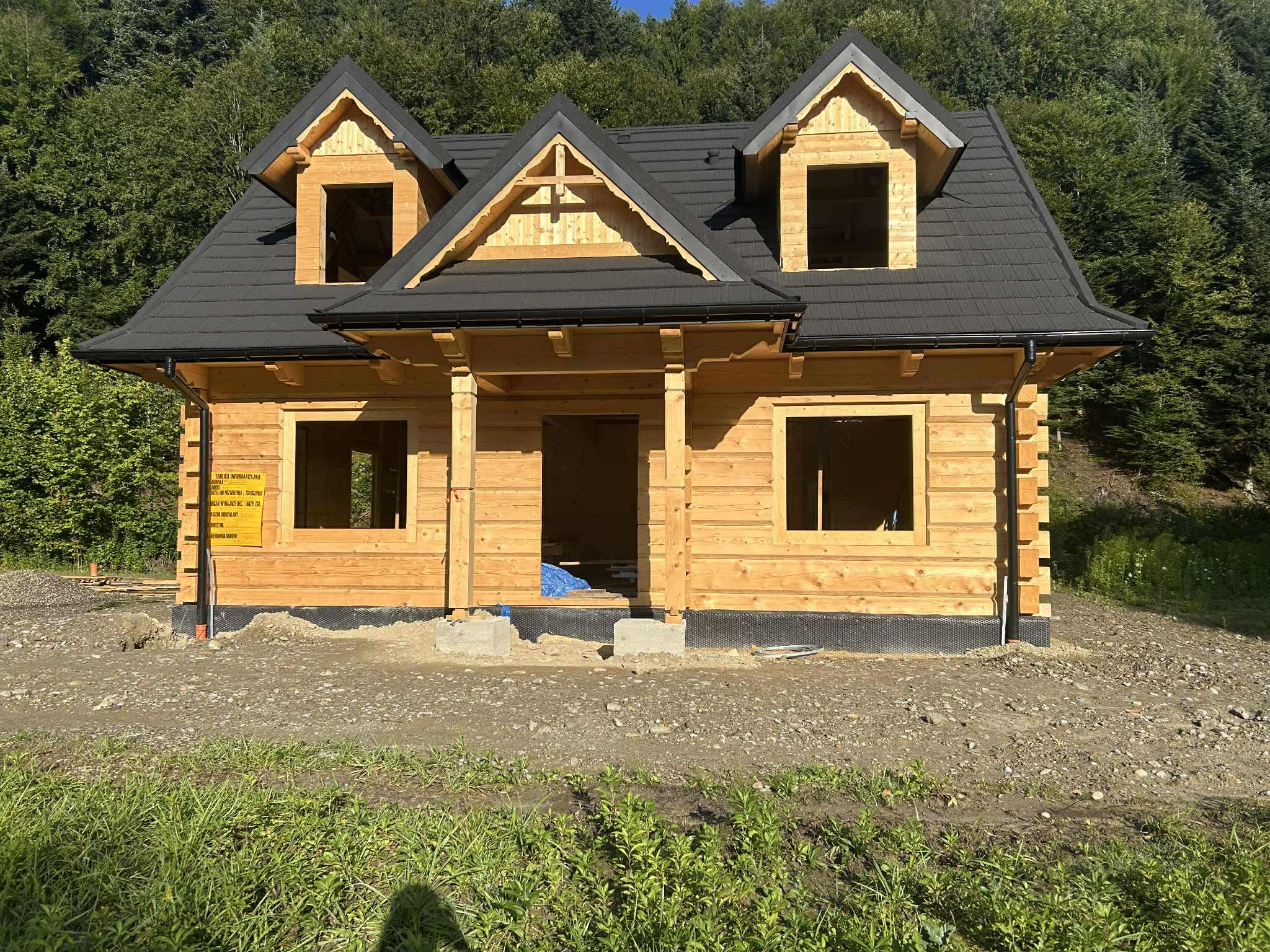 Niedokończony dom z bali z czarnym dachem na tle zieleni, widoczne detale konstrukcyjne i tablica informacyjna. Ujęcie z poziomu gruntu.