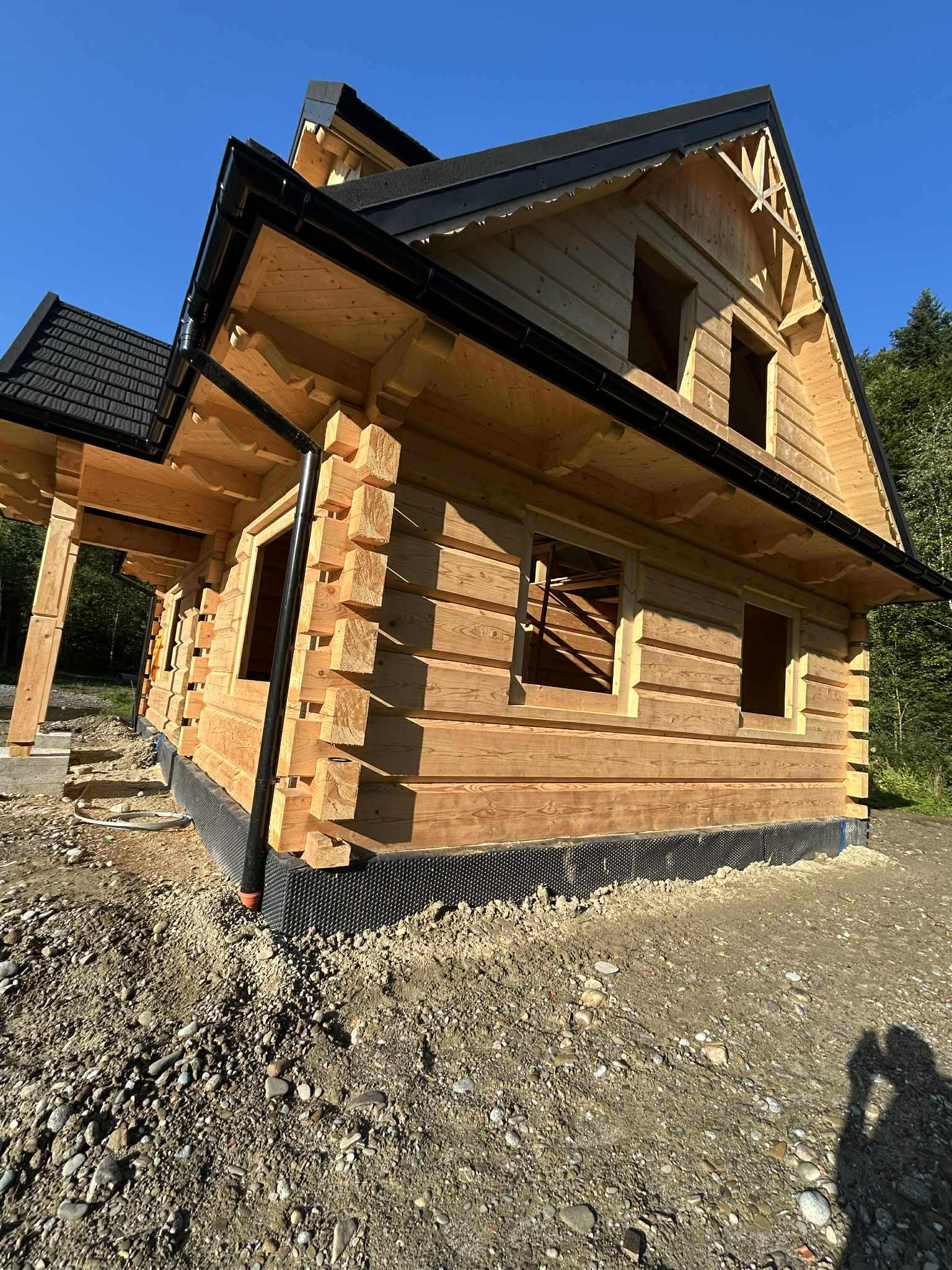 Surowy stan domu z bali z czarnym dachem i rynnami, widok z zewnątrz na tle zieleni. Widoczne detale konstrukcyjne i izolacja fundamentów.