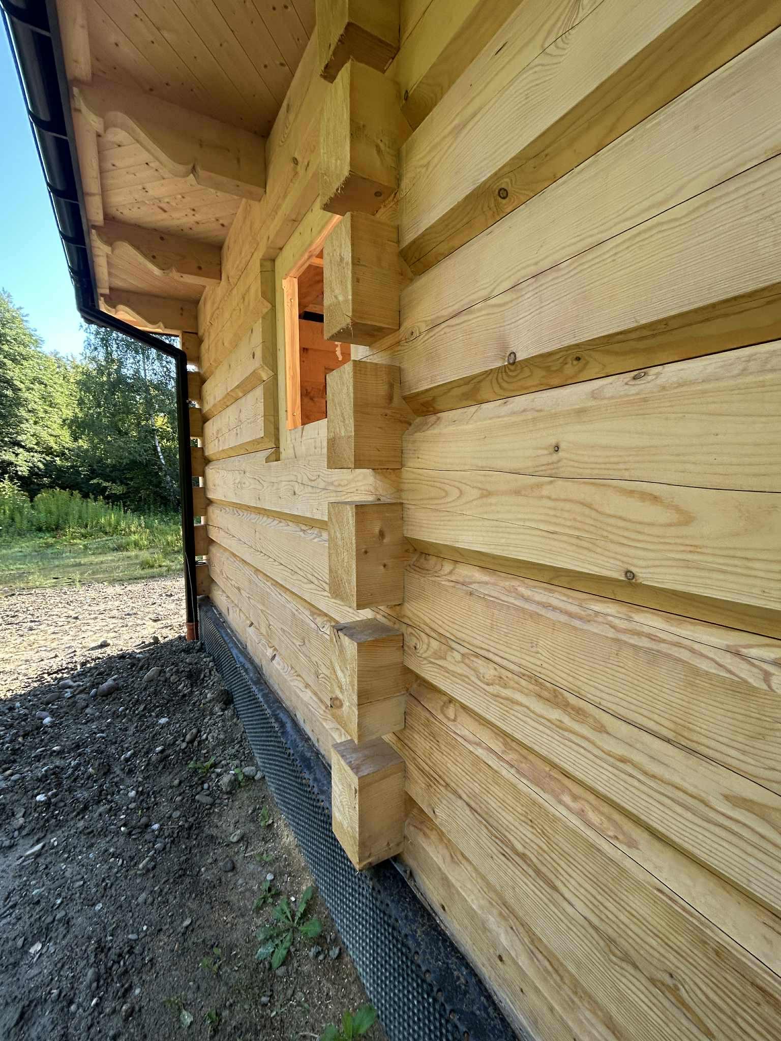 Fragment ściany zewnętrznej domu z bali, widoczny detal łączenia bali na rogu budynku, z czarną folią fundamentową przy ziemi. Surowe drewno, widoczne słoje.