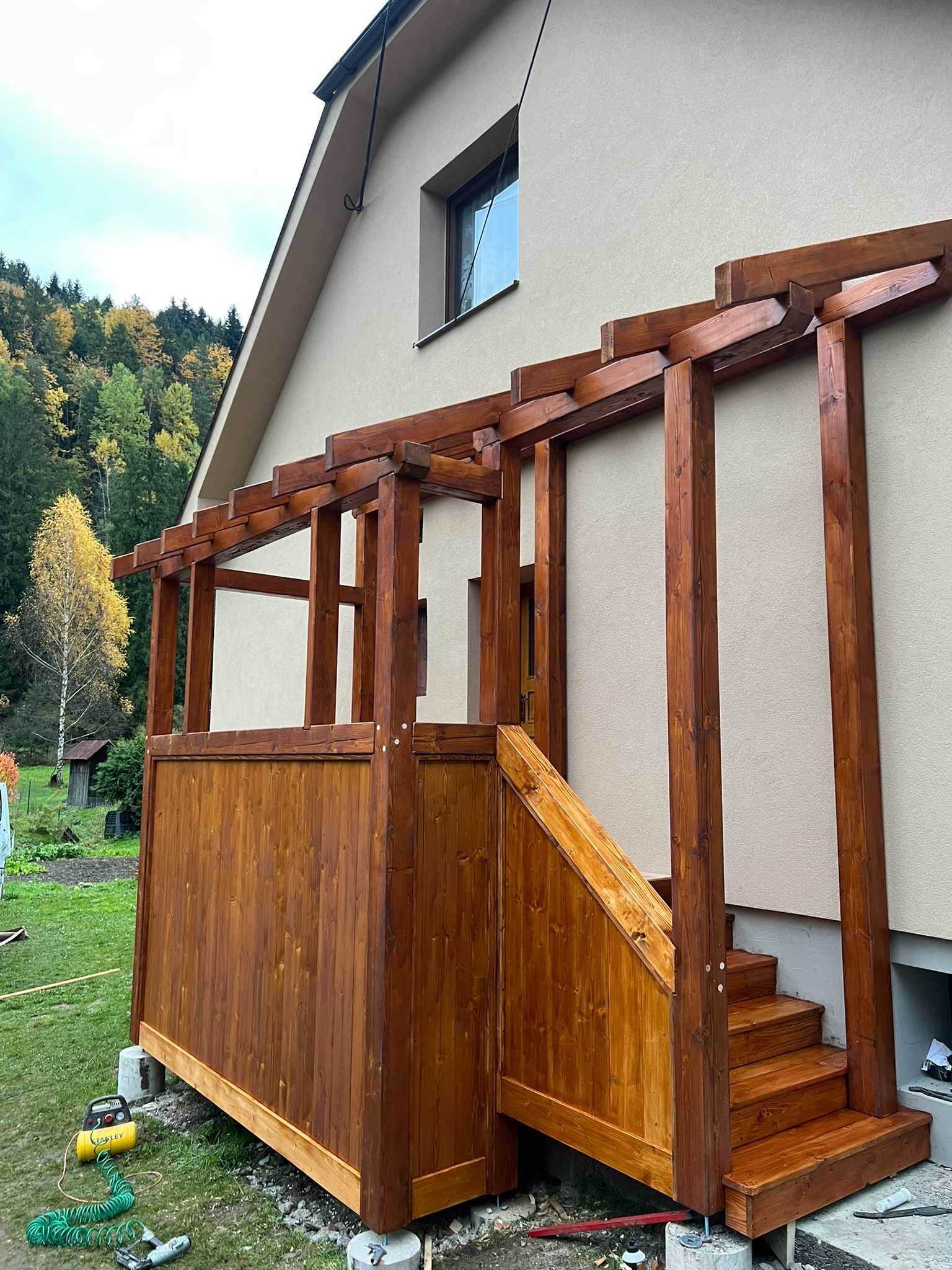 Drewniana altana z zadaszeniem, przylegająca do budynku z beżową elewacją, schody wejściowe. Na trawie narzędzia. Widok na las w tle.