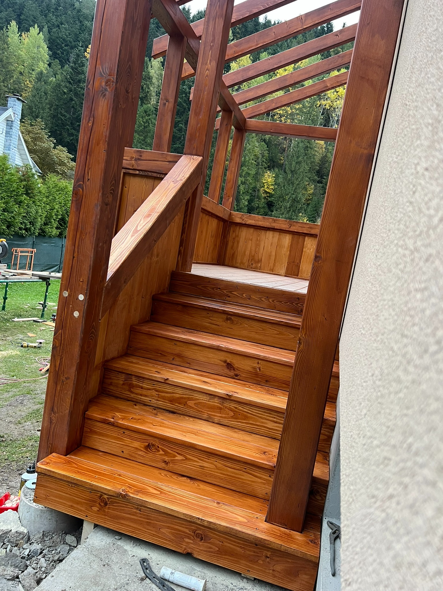 Drewniane schody z balustradą, prowadzące do drewnianej konstrukcji, prawdopodobnie werandy. Widoczne elementy montażowe i narzędzia w tle. Świeżo polakierowane drewno.