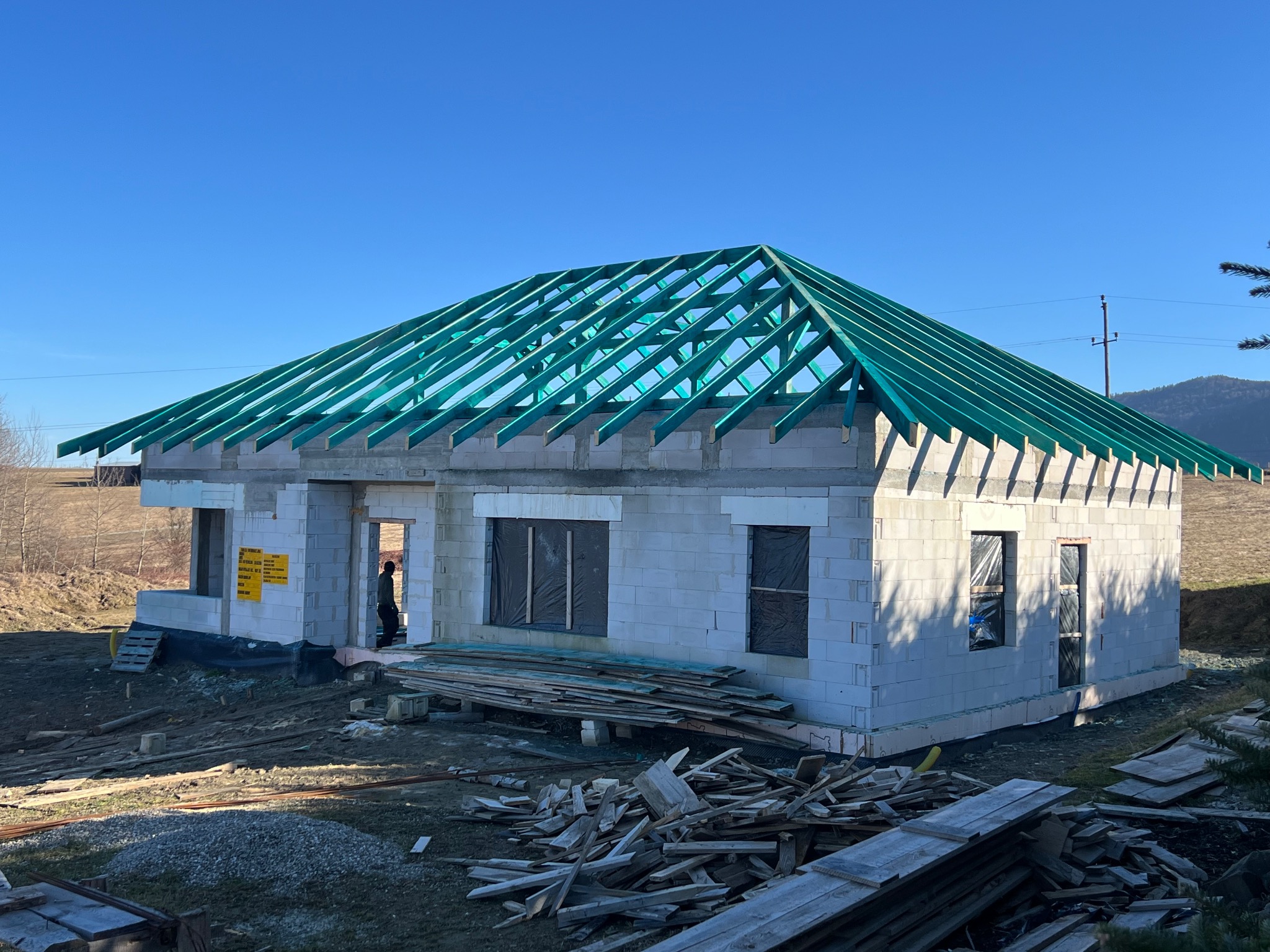 Surowy stan domu z betonu komórkowego z zieloną konstrukcją dachu na tle błękitnego nieba i pagórkowatego terenu. Widoczne deski i materiały budowlane.