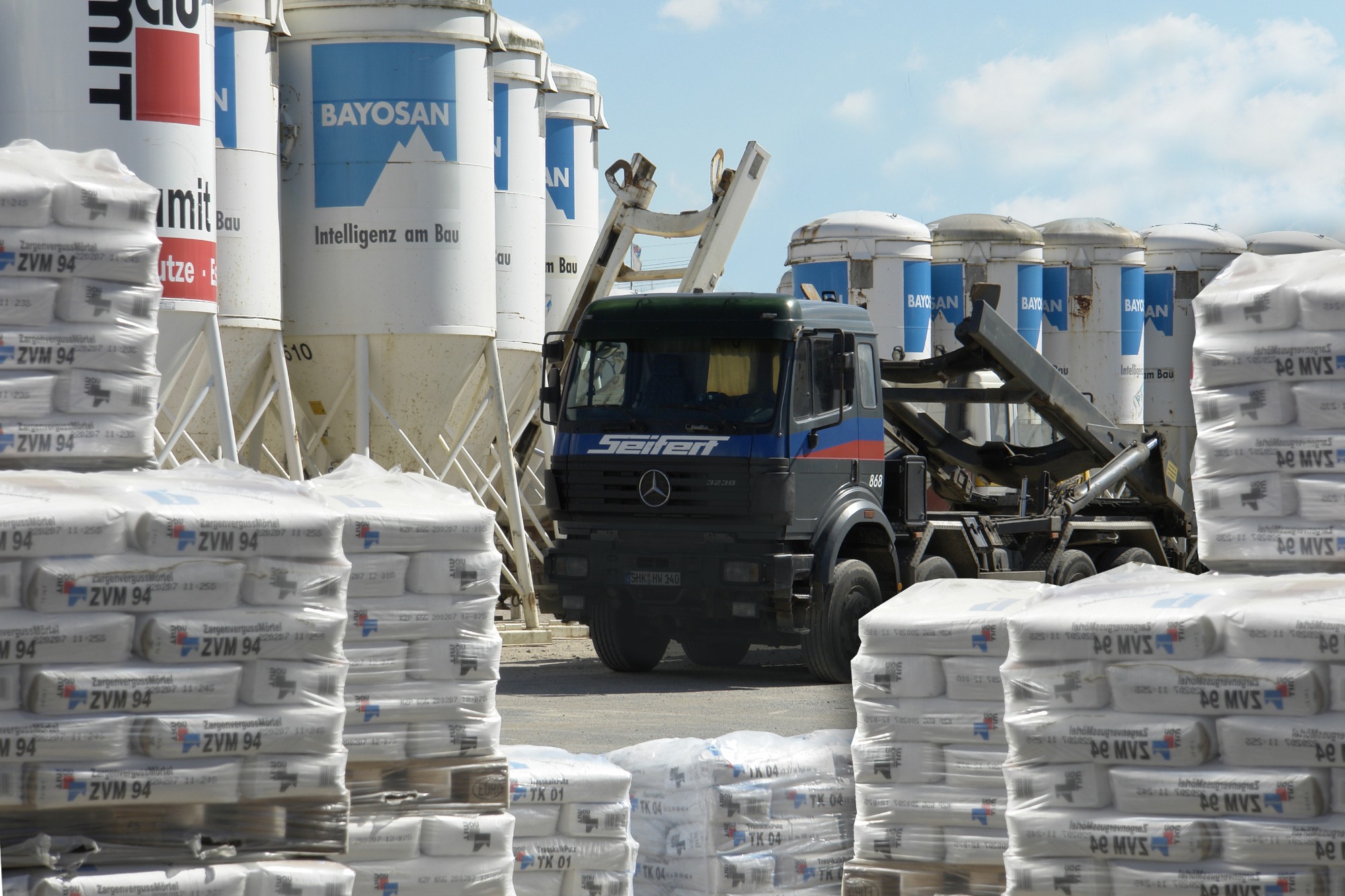 Ciężarówka Seifert z kontenerem hakowym na tle silosów Bayosan i palet z zapakowanym w folie cementem ZVM 94, widok z dołu.