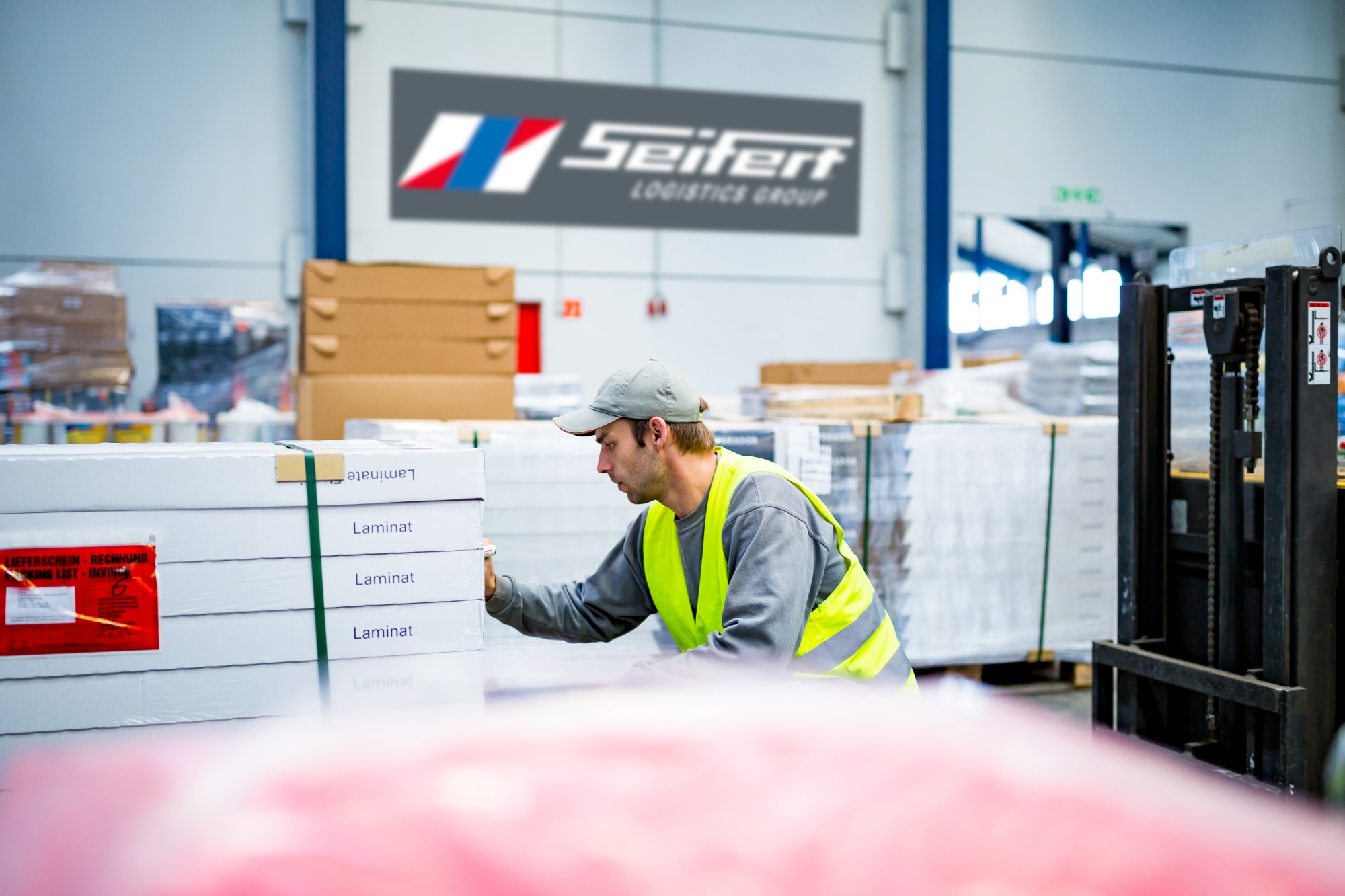 Pracownik w kamizelce odblaskowej sprawdza pakiet laminatu w magazynie z logo Seifert Logistics Group w tle, obok wózka widłowego.