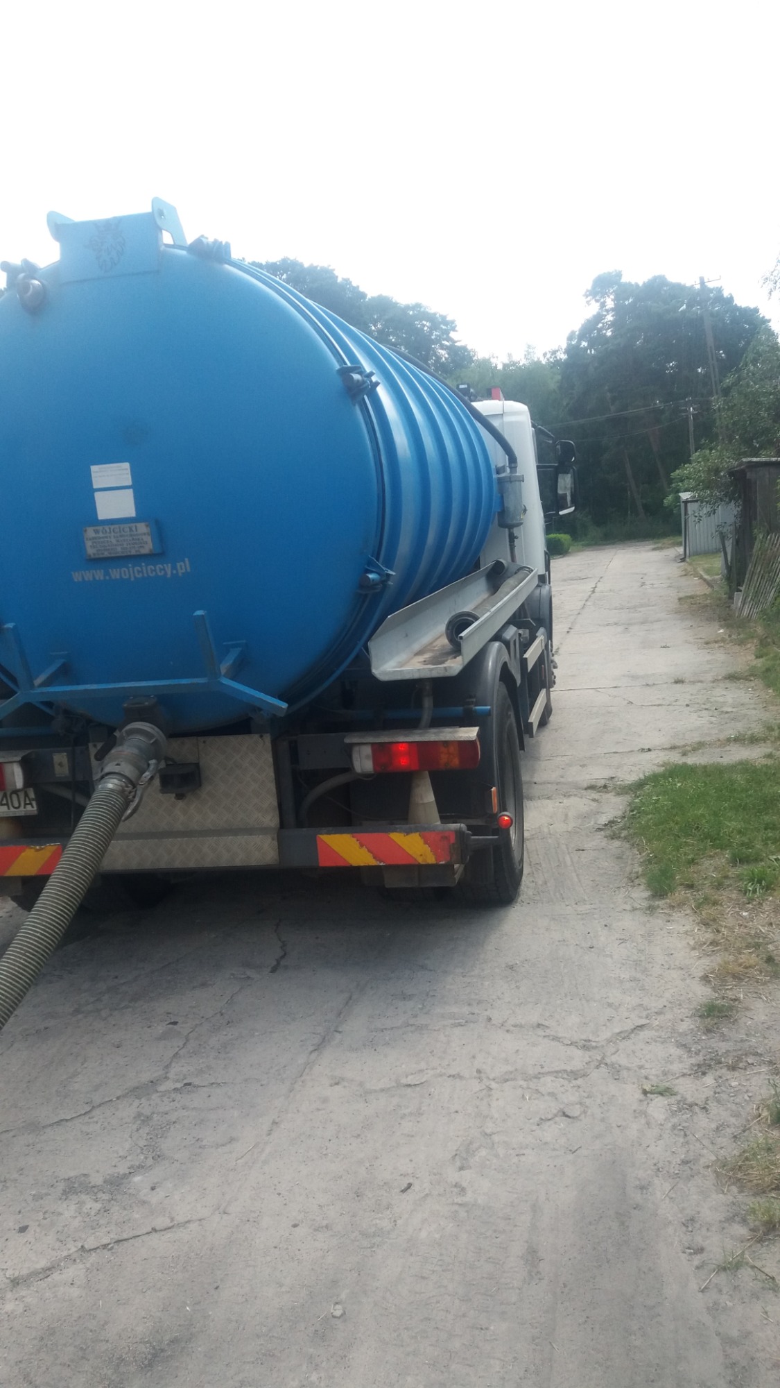 Ciężarówka asenizacyjna z niebieskim zbiornikiem, widok z tyłu, wąż podłączony, zaparkowana na szutrowej drodze.
