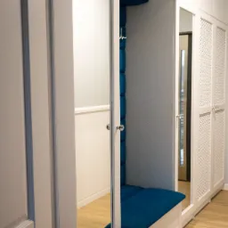 Biała szafa wnękowa z lustrzanymi drzwiami i siedziskiem z niebieską tapicerką, widok na wnętrze przedpokoju z drewnianą podłogą.