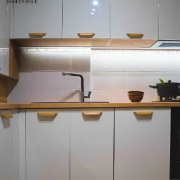 Biała kuchnia na wymiar z drewnianymi blatami i uchwytami, czarna bateria kuchenna, oświetlenie LED pod szafkami, logo firmy w rogu