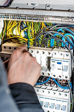 Elektryk w trakcie pracy nad skrzynką bezpiecznikową, widoczne liczne przewody w różnych kolorach oraz białe wyłączniki firmy Hager.