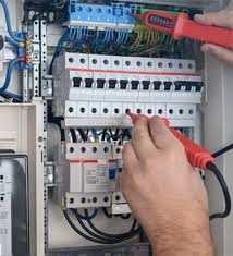 Elektryk sprawdza napięcie w rozdzielnicy elektrycznej za pomocą próbnika napięcia. Widoczne bezpieczniki, przewody i obudowa rozdzielnicy.