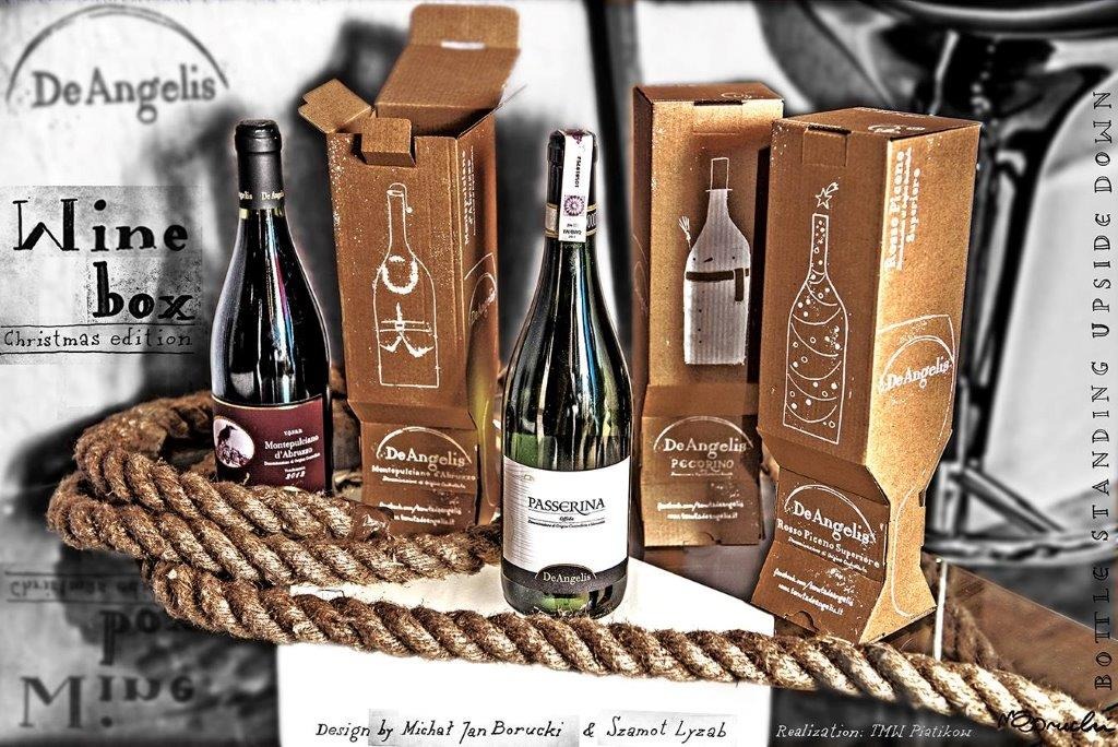 Dwie butelki wina stojące obok siebie, prezentowane na tle tekturowych pudełek z odręcznymi grafikami butelek i logiem De Angelis, całość ułożona na białym podłożu z grubym sznurem na pierwszym...