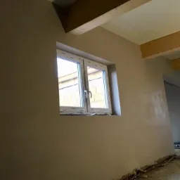 Wnętrze po nałożeniu gładzi, widoczne okno PCV i drewniane belki stropowe, ślady po pracach na podłodze.