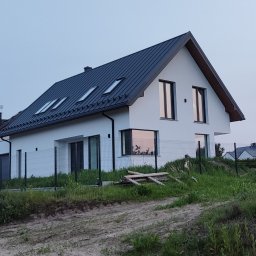 mijabodeSign - Nowoczesny, biały dom jednorodzinny z ciemnym dachem dwuspadowym i oknami dachowymi. Widok z zewnątrz, ogrodzenie siatkowe, trawa i ziemia na pierwszym planie.