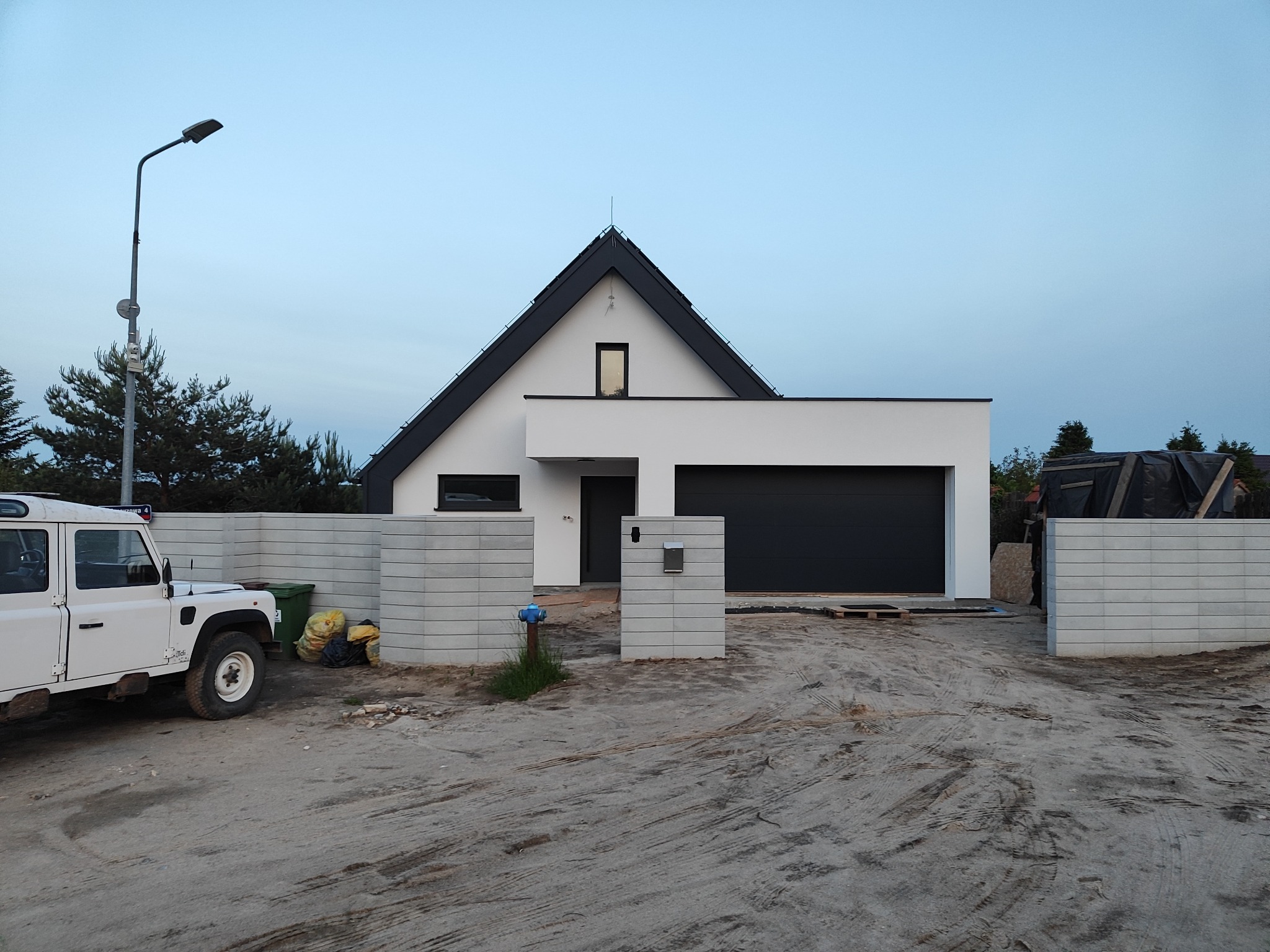 Nowoczesny dom jednorodzinny z garażem i elementami ogrodzenia z betonu architektonicznego, widziany z zewnątrz w trakcie budowy. Na pierwszym planie zaparkowany biały Land Rover.