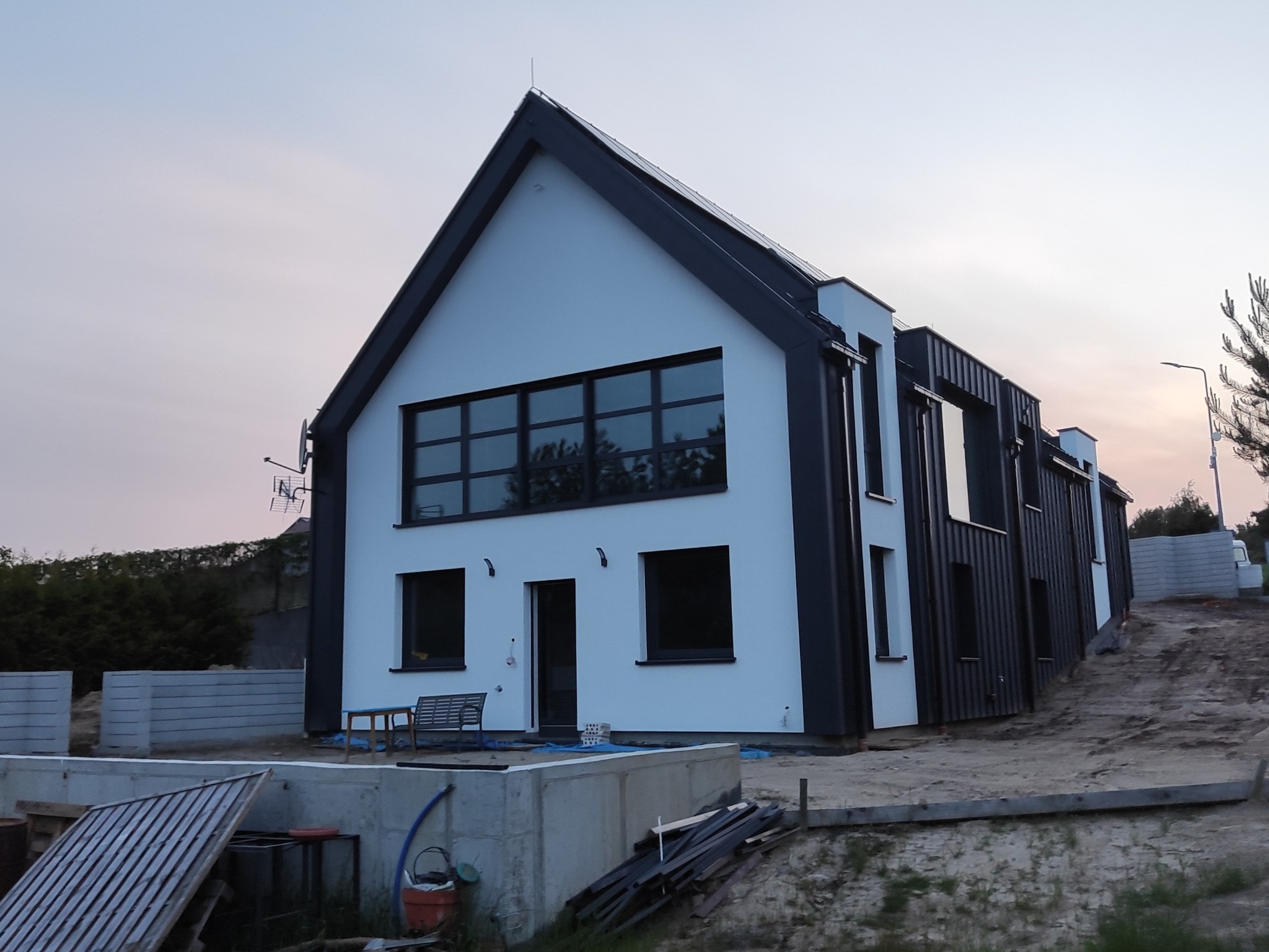 Nowoczesny dom jednorodzinny w trakcie budowy, z białymi ścianami i ciemną stolarką okienną. Widoczne fundamenty i elementy konstrukcyjne. Ujęcie z zewnątrz, o zmierzchu.