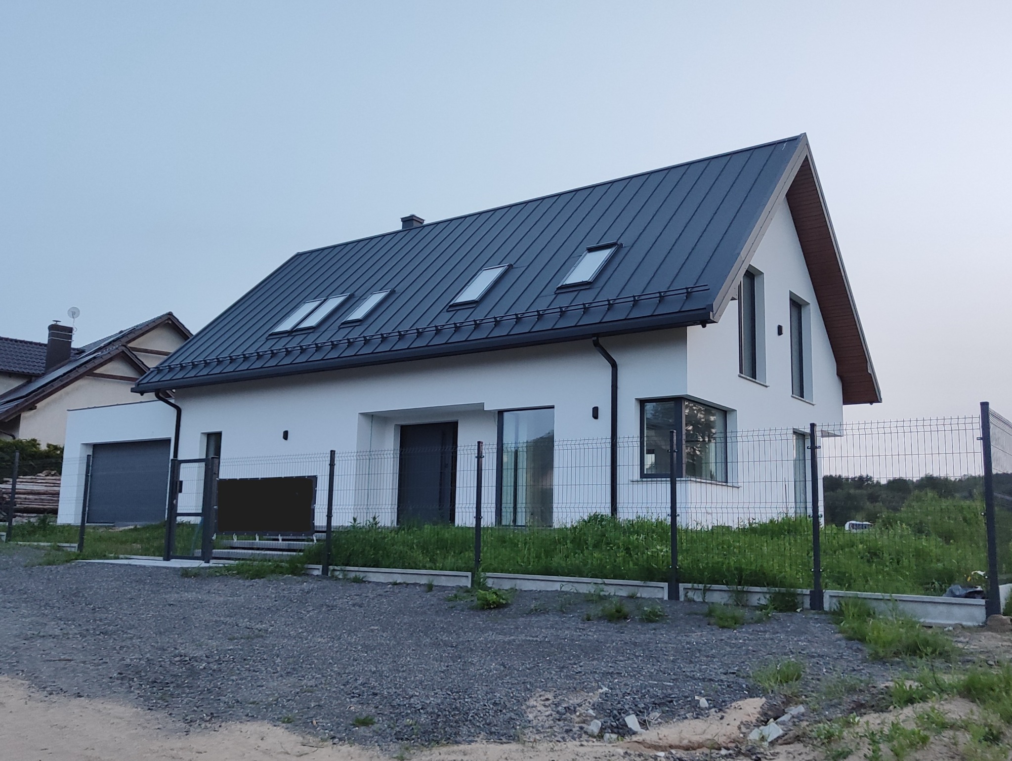 Nowoczesny, parterowy dom jednorodzinny z ciemną, skośną blachodachówką i trzema oknami dachowymi, otoczony metalowym ogrodzeniem panelowym.