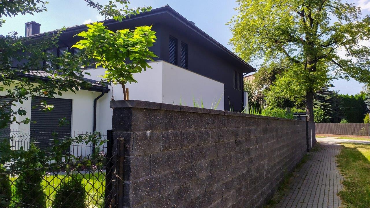 Dwukondygnacyjny dom jednorodzinny z ciemną elewacją i białymi elementami, widoczny zza betonowego muru, z młodym drzewkiem zasadzonym na jednym z filarów muru.