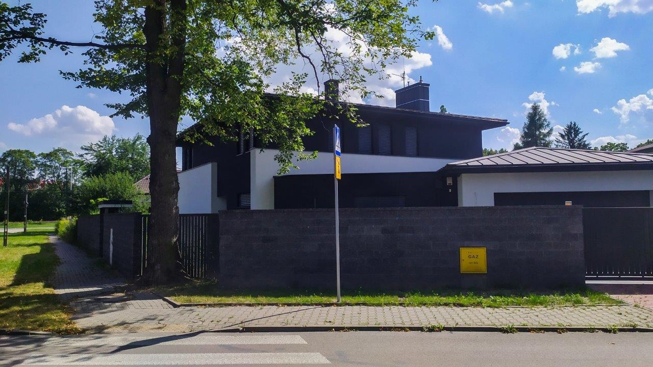 Nowoczesny dom jednorodzinny w Redzie z czarno-białą elewacją, widoczny zza ceglanego muru i drzewa, w tle błękitne niebo.