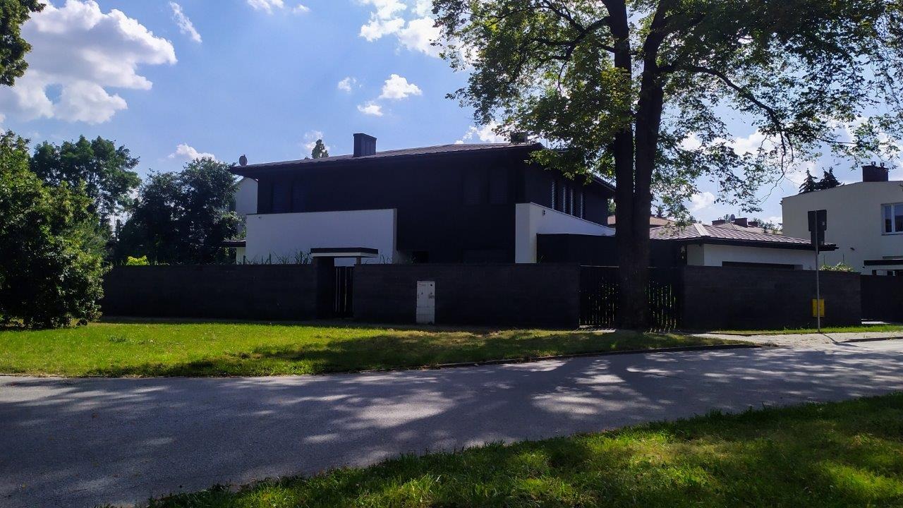 Nowoczesny dom jednorodzinny z ciemną elewacją i białymi akcentami, otoczony zielonym trawnikiem i wysokim ciemnym ogrodzeniem, widziany z perspektywy ulicy w słoneczny dzień.