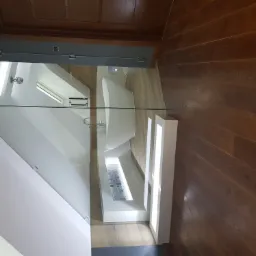 Nowoczesna łazienka z wolnostojącą białą wanną widoczna przez szklaną ścianę prysznica, drewniana podłoga w odcieniach brązu.