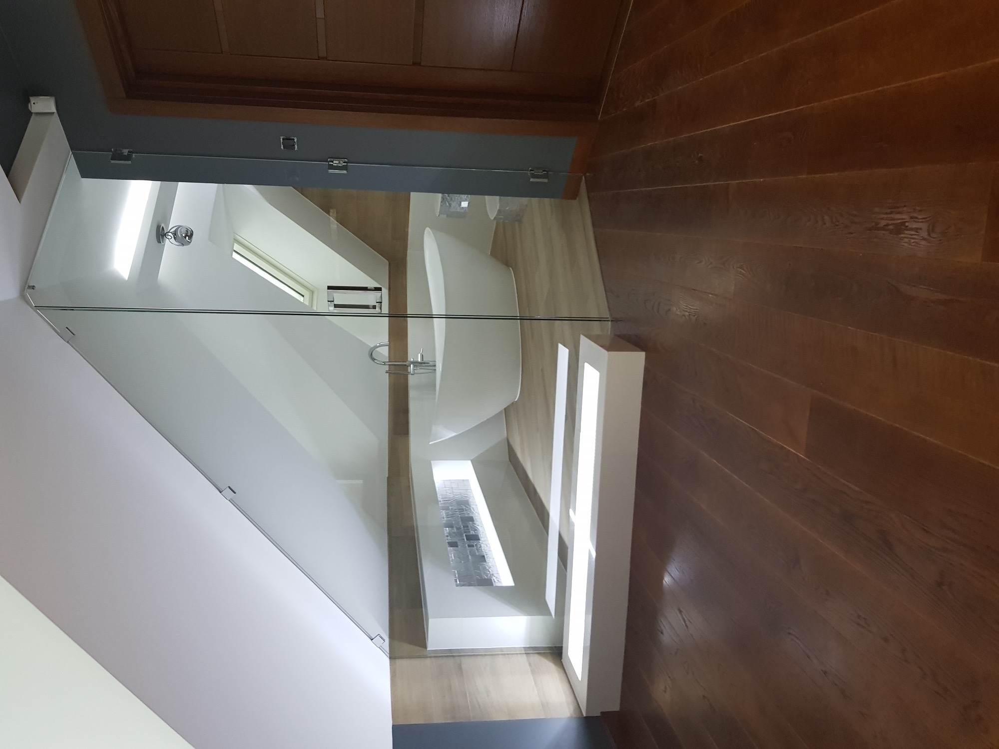 Nowoczesna łazienka z wolnostojącą białą wanną widoczna przez szklaną ścianę prysznica, drewniana podłoga w odcieniach brązu.