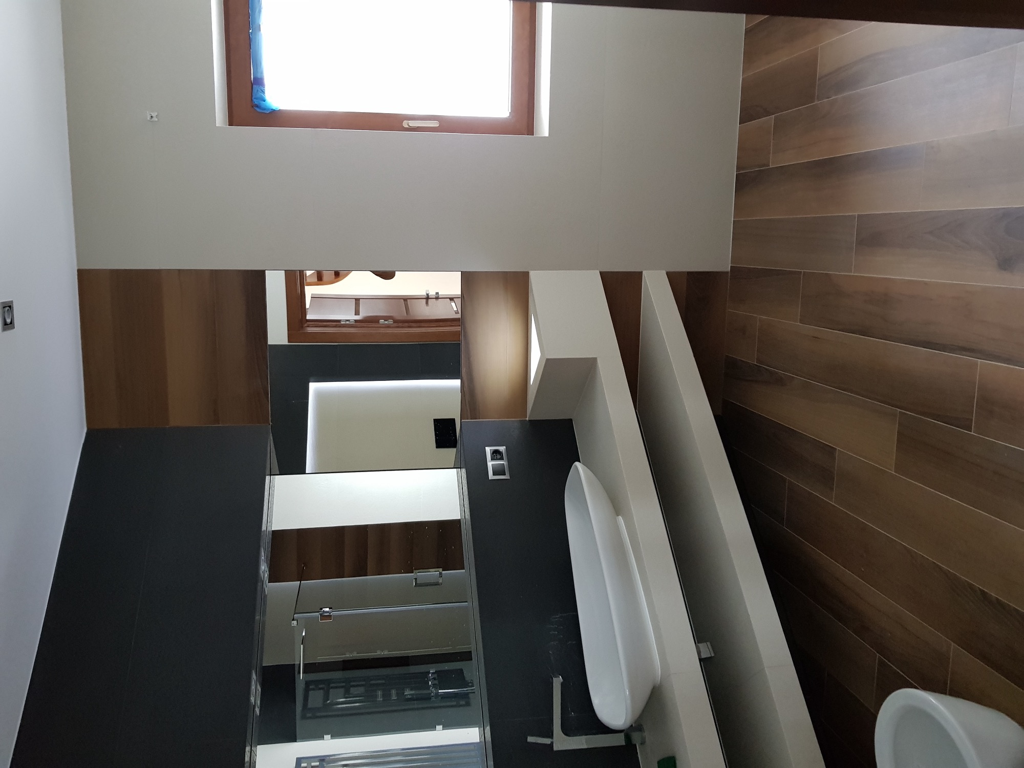 Nowoczesna łazienka po remoncie z drewnopodobnymi płytkami na ścianie, białą ceramiką i szarą zabudową meblową, okno zabezpieczone folią malarską.