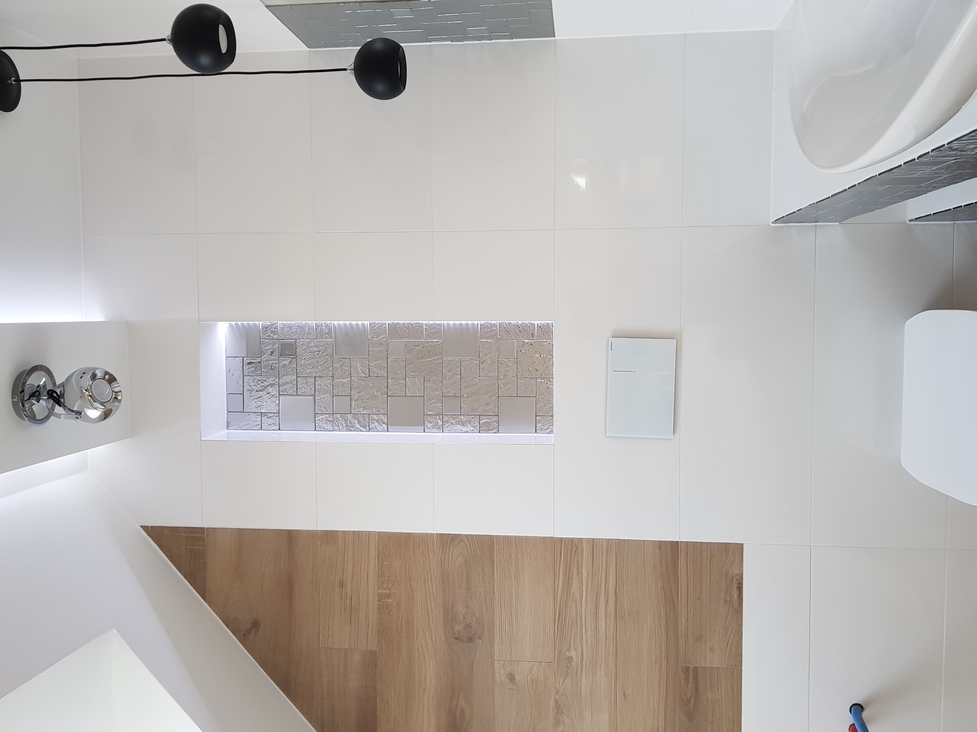 Nowoczesna łazienka z białymi płytkami, drewnianą okładziną, podświetlaną wnęką z mozaiką i minimalistycznym oświetleniem sufitowym.