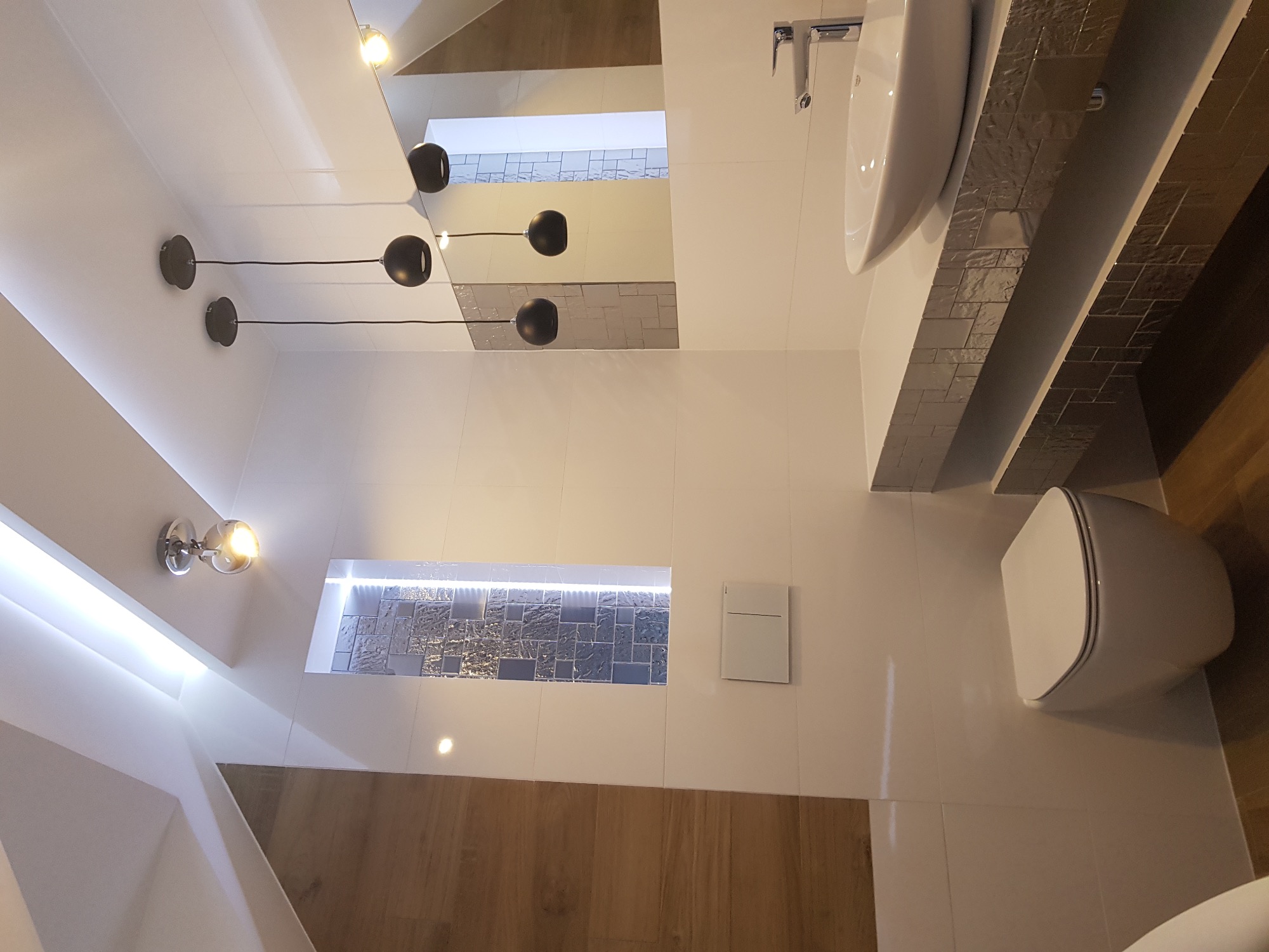 Nowoczesna łazienka z białymi płytkami, podwieszaną toaletą i umywalką, oświetlona lampami sufitowymi i kinkietem, z dekoracyjną wnęką wyłożoną mozaiką.