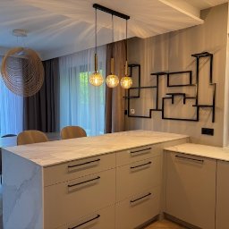 Anna Zakrzewska Projektowanie Wnętrz - Nowoczesna kuchnia z wyspą, marmurowym blatem i designerskim oświetleniem. Na ścianie oryginalna, metalowa dekoracja. Widok na jadalnię i okno z roletą rzymską.